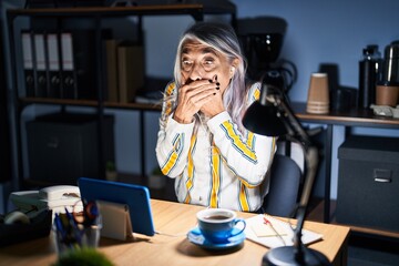 Middle age woman with grey hair working at the office at night shocked covering mouth with hands for mistake. secret concept.