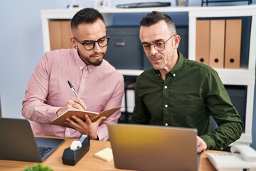 Two men business workers using laptop writing on notebook at office