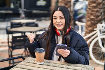 Young hispanic woman using smartphone sitting on the table pointing thumb up to the side smiling happy with open mouth