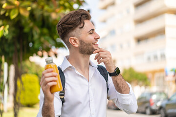 Young handsome man holding an orange juice at outdoors thinking an idea and looking side