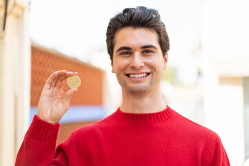Young handsome man holding a Bitcoin at outdoors smiling a lot