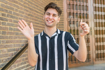 Naklejka premium Young handsome man holding home keys at outdoors saluting with hand with happy expression