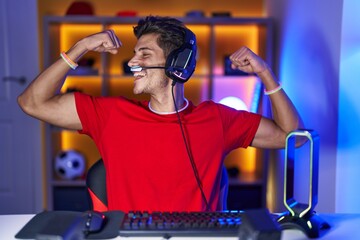 Young hispanic man playing video games showing arms muscles smiling proud. fitness concept. - obrazy, fototapety, plakaty