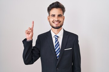 Young hispanic man with tattoos wearing business suit and tie showing and pointing up with finger number one while smiling confident and happy.