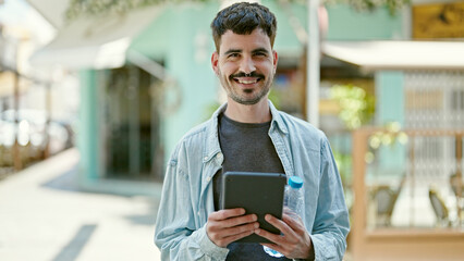 Young hispanic man smiling confident using touchpad holding bottle of water at coffee shop terrace