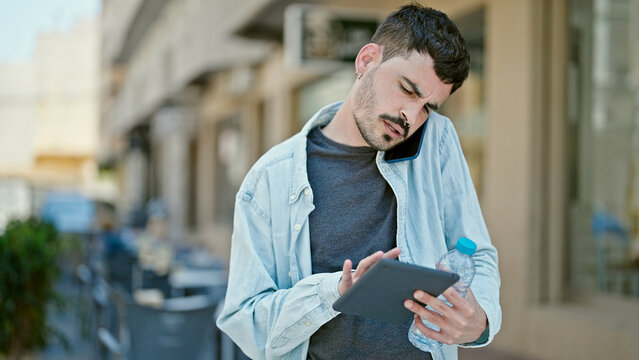 Young hispanic man using touchpad talking on smartphone holding bottle of water at coffee shop terrace