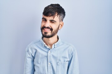 Fototapeta premium Young hispanic man with beard standing over blue background winking looking at the camera with sexy expression, cheerful and happy face.