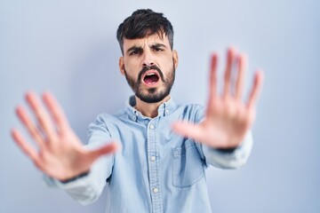 Young hispanic man with beard standing over blue background doing stop gesture with hands palms,...
