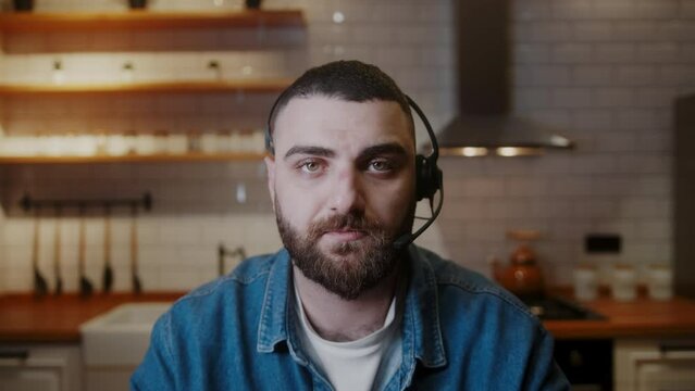 Bearded young adult man wears wireless headset with microphone talking to camera making conducting conference business call in kitchen. Having video call zoom meeting