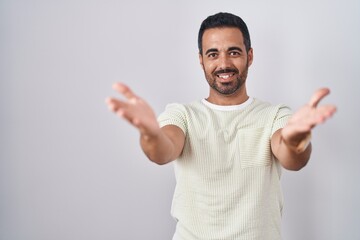 Fototapeta premium Hispanic man with beard standing over isolated background smiling cheerful offering hands giving assistance and acceptance.