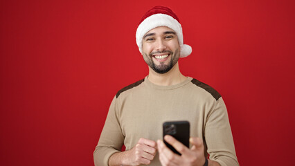 Young arab man wearing christmas hat using smartphone over isolated red background