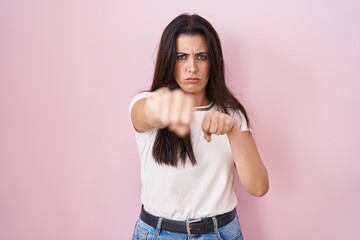 Young brunette woman standing over pink background punching fist to fight, aggressive and angry attack, threat and violence