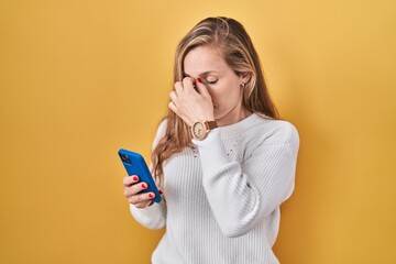 Young blonde woman using smartphone typing message tired rubbing nose and eyes feeling fatigue and...