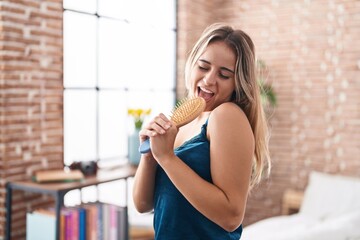 Obraz premium Young blonde woman singing song using brush as a microphone at bedroom