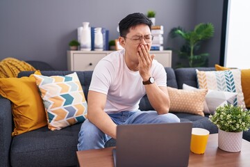 Young asian man using laptop at home sitting on the sofa bored yawning tired covering mouth with...