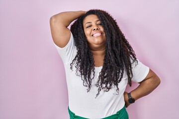 Plus size hispanic woman standing over pink background smiling confident touching hair with hand up...