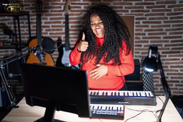 Plus size hispanic woman playing piano at music studio doing happy thumbs up gesture with hand. approving expression looking at the camera showing success.