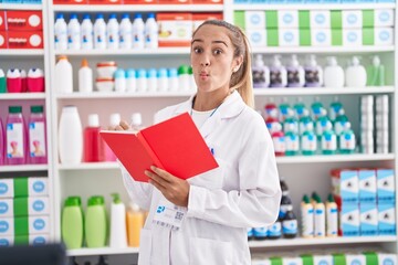 Young blonde woman working at pharmacy drugstore holding notebook making fish face with mouth and squinting eyes, crazy and comical.