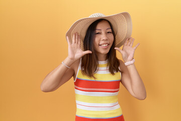 Middle age chinese woman wearing summer hat over yellow background smiling cheerful playing peek a...