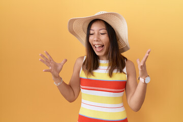 Middle age chinese woman wearing summer hat over yellow background celebrating mad and crazy for success with arms raised and closed eyes screaming excited. winner concept