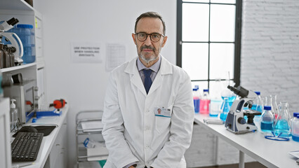 Serious-faced, middle-aged scientist with grey hair, sitting amid the buzz of his lab, immersed in...