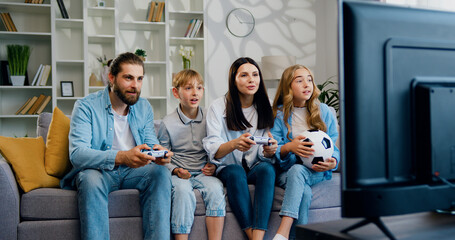 Happy smiling family of mother, father, son, and daughter playing video game together sitting on a couch at home. Parents play online using a controller and children cheer. Relationship, bonding - obrazy, fototapety, plakaty