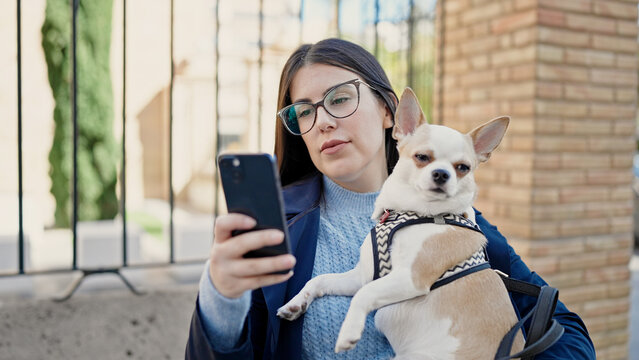 Young hispanic woman with chihuahua dog standing with serious expression taking a selfie picture at street