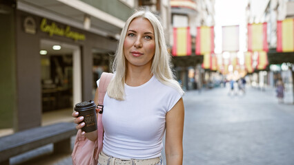 Relaxed and beautiful young blonde woman holding a hot take away coffee cup on a sunny city street, expressing her cool urban lifestyle outdoors.