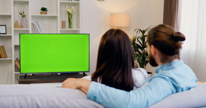 Couple sitting together on couch at home, watching tv shows with chroma-key green screen. Wife and husband resting at home together. Green screen. Chromakey. Home theater.