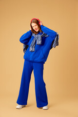 Fototapeta premium Full-length image of young girl in stylish blue clothes and shirt, listening to music in headphones against beige studio background. Concept of youth culture, fashion, human emotions, lifestyle, music