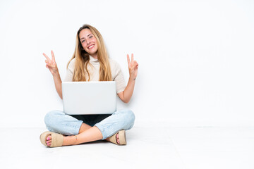 Young woman with laptop sitting on the floor isolated on white background showing victory sign with...