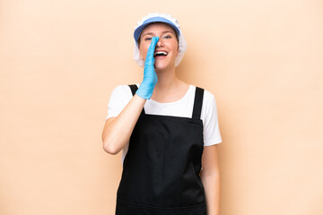 Fishmonger caucasian woman wearing an apron and holding a raw fish isolated on beige background shouting with mouth wide open