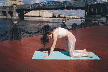Fototapeta na wymiar A woman in sportswear performs a cat-cow stretch on a yoga mat placed on a red wooden deck, with a scenic bridge and river in the background