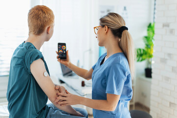 Diabetologist doctor teaching young patient ho to connect continuous glucose monitor sensor to smartphone, checking teenage boy's glucose data in real time. Endocrinologist taking care of teenage - obrazy, fototapety, plakaty