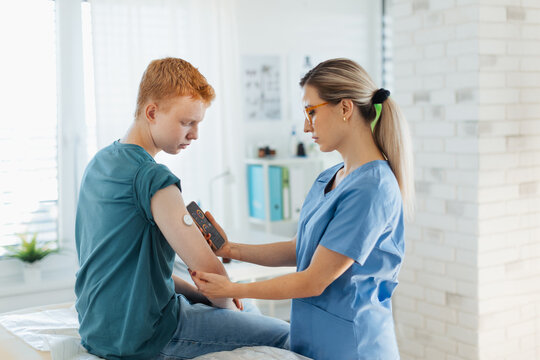 Diabetologist doctor connecting continuous glucose monitor sensor to smartphone, checking teenage boy's glucose data in real time. Endocrinologist taking care of teenage diabetic patient.