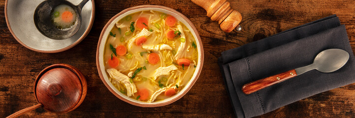 Chicken noodle soup with vegetables panorama, a bowl of healthy broth, overhead flat lay shot on a rustic wooden table, winter home cooking