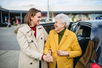 Senior lady getting out of the car, caregiver helping her, holding her hands. Elderly woman has problem with standing up from the car back seat. - obrazy, fototapety, plakaty