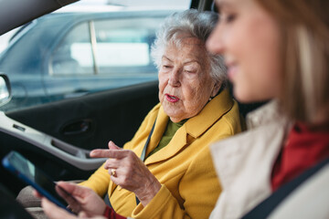 Granddaughter driving her elderly grandmother in the car, taking her to the doctor, shopping or to the bank. Caregiver driving elderly lady to the church, pharmacy, searching for route on a smartphone - obrazy, fototapety, plakaty
