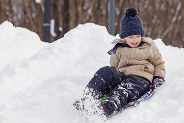 Happy boy slides down an ice slide in winter outside. A little boy rides downhill on a sled in...