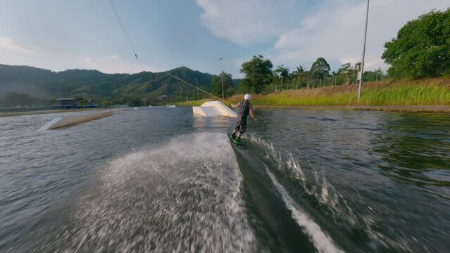 Aerial view sport man riding wakeboarding jibbing rotation trick track ramps tropical sunny seascape. FPV sport drone follow low shot active extreme male enjoy speed splash ride wakeboard cable jump