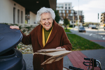 Elderly woman disposing cardboard into the garbage can, waste container in front of her apartment complex.