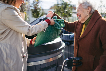 Woman helping elderly neighbor throw away trash into garbage can, waste container in front of their...