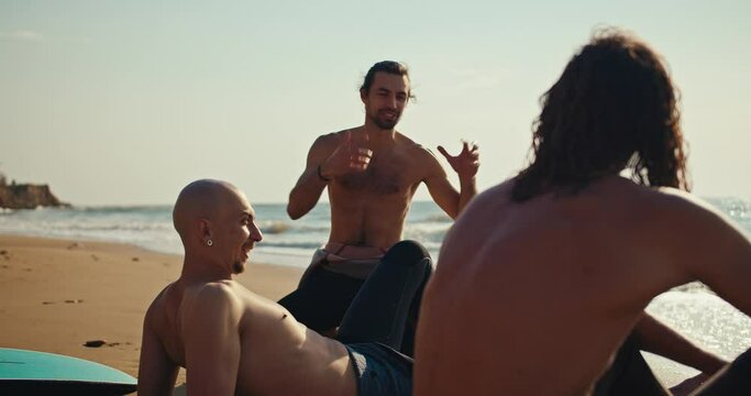A brunette man with a naked torso talks about his emotions surfing to his friends by a surfer in wetsuits on a sandy beach near the sea
