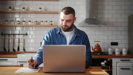 Bearded young adult man wearing in-ear headphones typing on laptop doing research sitting against the kitchen working on laptop  - Powered by Adobe