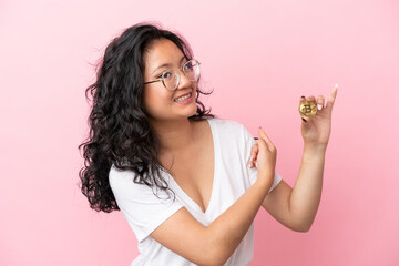 Young asian woman holding a Bitcoin isolated on pink background pointing back