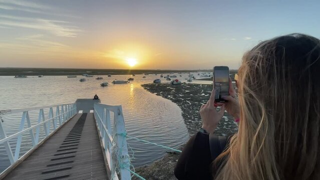 Mulher filmando e fotografando o por do sol em uma doca de barcos, em frente ao mar de Faro - Portugal. 