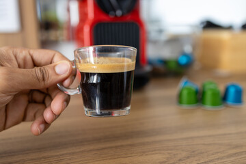 brown hand holding a fresh espresso in a glass mug.
