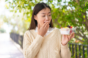 Young Chinese woman holding a bowl of fruit at outdoors with surprise and shocked facial expression