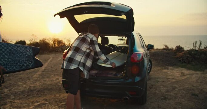 A guy in a plaid shirt and a green hat, his friend in a blue sweatshirt and floating in a white shirt are putting their surfboards in the open trunk of a black car at Sunrise in the morning