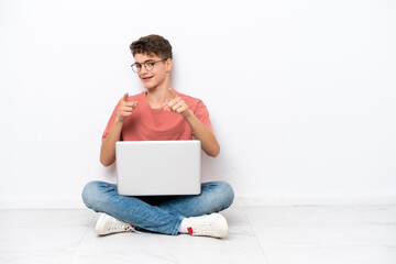Teenager Russian man holding pc sitting on the floor isolated on white background pointing to the...